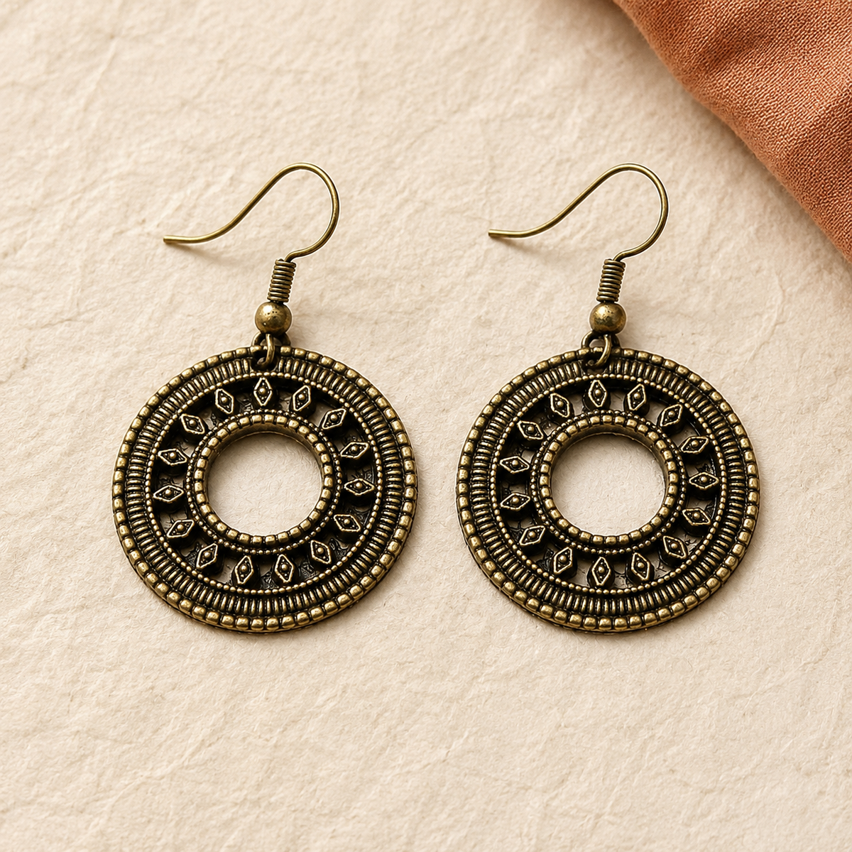 A pair of round drop earrings with intricate filigree detailing in an antique bronze finish, displayed on a textured beige surface with a piece of rusted orange fabric in the corner.