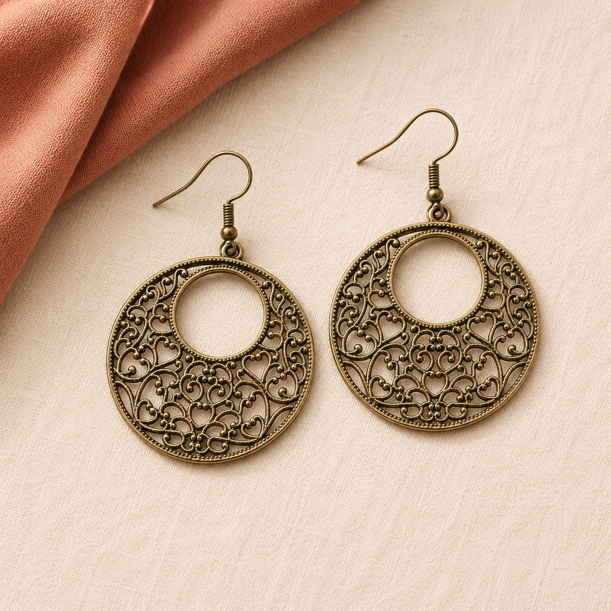 A pair of antique bronze filigree hoop earrings with fishhook wires, displayed on a textured cream surface next to a draped piece of peach fabric.