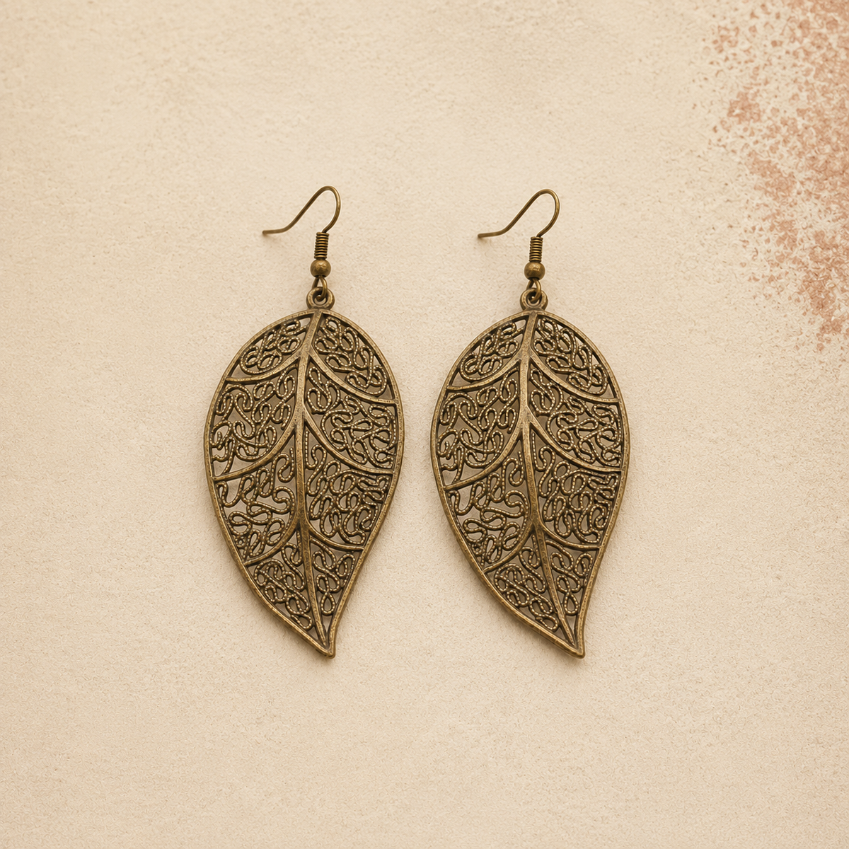 A pair of antique bronze leaf-shaped dangle earrings with intricate filigree cutouts and fishhook wires, displayed on a textured beige background.