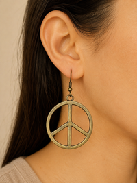 A close-up of a model's ear wearing a large bronze peace sign dangle earring with a hook back.