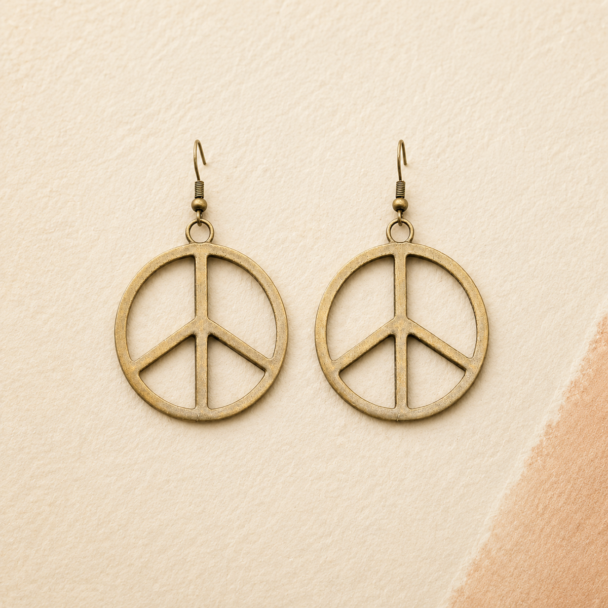 A pair of round peace sign dangle earrings in an antique bronze finish, displayed on a textured beige background with a soft peach-colored corner accent.