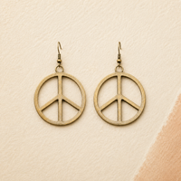 A pair of round peace sign dangle earrings in an antique bronze finish, displayed on a textured beige background with a soft peach-colored corner accent.