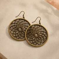 A pair of round, antique bronze-tone dangle earrings with intricate web-like filigree patterns, displayed on a textured beige surface.